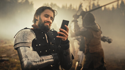 Portrait of Handsome Medieval Knight Using Smartphone on Battlefield, Smiling. Funny Concept: Armored Warrior Having Fun, Ordering Online, Betting, Investing, doing E-commerce. War is Raging - obrazy, fototapety, plakaty