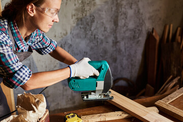 Female Carpenter sawing wood with an electric jigsaw. Woman in apron doing some carpentry work in...