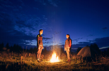 Horizontal snapshot of couple of tourists spending time together in camping. Young boy and girl near campfire in the evening outdoors in nature. Standing looking at fire in summer night