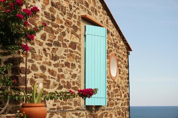 Fototapeta na wymiar Gable wall of stone house featuring turquoise coloured wooden window shuuters and plant with red flowers in bloom with Mediterranean sea visible against backdrop of blue sky, Collioure, France