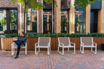 Millennial man in trousers and black jacket is sitting on one of four chairs in courtyard of modern brick residential complex with landscaping on city street