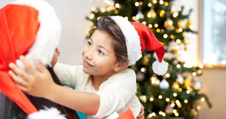 winter holidays, family and people concept - happy mother and little girl in santa hats at home over christmas tree background
