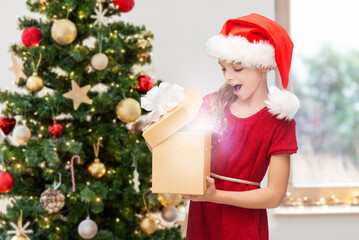 Obraz na płótnie Canvas winter holidays and celebration concept - happy surprised girl opening gift box with fairy dust at home over christmas tree background