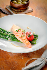 Baked salmon steak on a white plate with asparagus, cherry tomatoes and sauce. Salmon fillet dish served on a wooden table