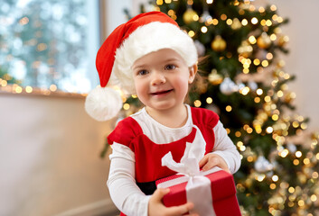 christmas, holidays and childhood concept - happy baby girl in red dress and santa helper hat holding gift at home