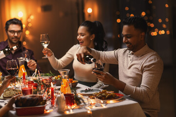 holidays, party and celebration concept - multiethnic group of happy friends having christmas dinner at home and drinking red wine