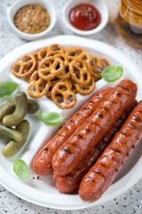 White disposable plate with grilled bratwurst sausages, pickles, mini pretzels, dips and beer, close-up, vertical shot, selective focus