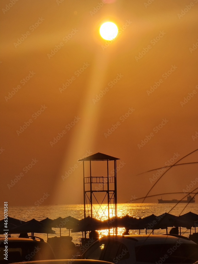 Sticker Vertical shot of a seacoast with umbrellas and security tower under an orange sky at sunset