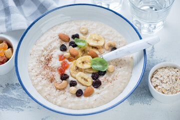 Plate of oatmeal porridge topped with dried banana, candied fruits and nuts, horizontal shot on a white concrete surface