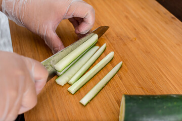 Julienning a cucumber into long thin strips on a wooden cutting board while wearing plastic gloves.