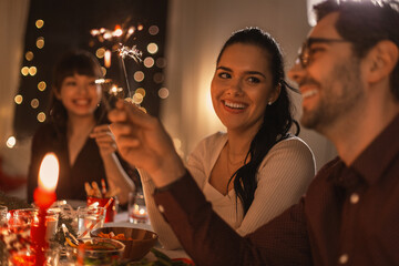 holidays and celebration concept - multiethnic group of happy friends with sparklers having christmas dinner at home