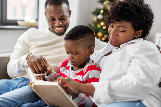 Family, Winter Holidays And People Concept - Happy African American Mother, Father And Baby Son Reading Book At Home On Christmas