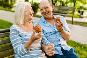 Fototapeta premium White senior couple smiling and eating muffins while sitting on bench