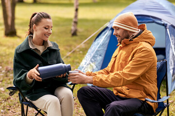 Naklejka na ściany i meble camping, tourism and travel concept - happy couple with thermos pouring tea to cup at tent camp