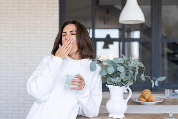 Young woman in a warm white hoodie at home in the kitchen, starts her day with a cup of coffee,...