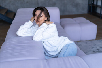A young woman at home in a white hoodie on the couch sits peacefully, enjoys loneliness, comfort, alone, in a calm mood