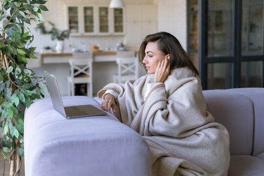 Young Woman At Home On The Couch In Winter Under A Cozy Blanket With A Laptop, Making Video Calls To Family Friends, Working Remotely