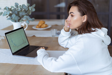 Young woman at home in the kitchen in a white hoodie with a laptop, advisor financial business...