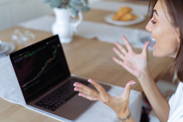 Young woman at home in the kitchen in a white hoodie with a laptop, a graph of growth on the screen, rejoices the cryptocurrency is growing up