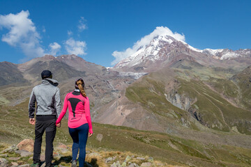 Naklejka premium A couple enjoying the cloudless view on Mount Kazbeg in Caucasus, Georgia. There slopes are barren and stony below the snow-capped peak and the Gergeti Glacier. Tranquillity. Natural remedy. Adventure