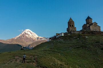 A man enjoying the view on Gergeti Trinity Church in Stepansminda, Georgia. The church is located on a high Caucasian mountain. Clear and blue sky above the church. In the back there are high mountain