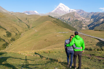Obraz premium A couple enjoying the cloudless view on Mount Kazbeg in Caucasus, Georgia. There slopes are barren and stony below the snow-capped peak and the Gergeti Glacier. Tranquillity. Natural remedy. Adventure