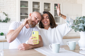 Obraz na płótnie Canvas Young couple husband with wife at home in the kitchen, happy chatting by video call, taking selfie on mobile phone