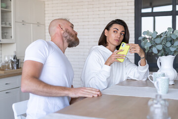 Young couple husband with wife at home in the kitchen, woman hides the phone screen, man tries to peek, distrust in the family