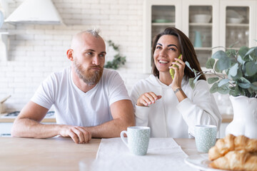 Young couple husband and wife at home in the kitchen, the girl cheerfully talks on the phone, the man sits unhappy with a bored look