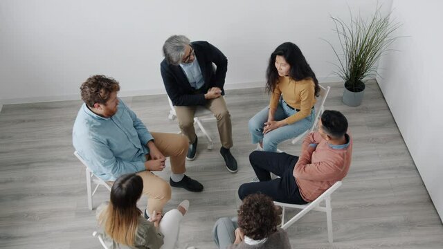 Asian woman is sharing difficult experience during support group meeting then doing high-five with caring man. People are sitting in circle inside beautiful studio.