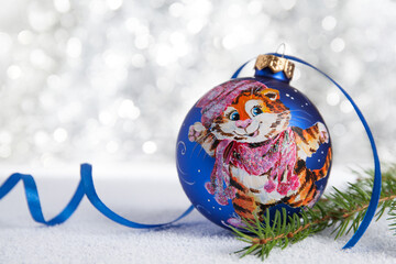 Christmas decoration - a hand-painted ball with the symbol of the Year of the Tiger on the skates. Nearby is a New Year tree branch and a blue ribbon. Blurred background of silver color