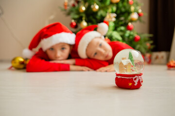 two children, brother and sister in red Christmas pajamas look at a glass ball with a Christmas tree and a house inside, dream and make wishes. new Year's winter concept. celebrating at home