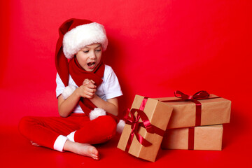 Merry Christmas. Portrait of a cute boy in a santa cap with gift boxes on a red background. A place for text.