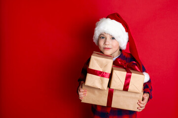 Happy holiday and Merry Christmas. Portrait of a boy in a cap with gifts in his hands on a red background. A place for text.
