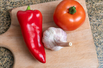 There is a tomato, red pepper, a head of garlic on a wooden kitchen plate