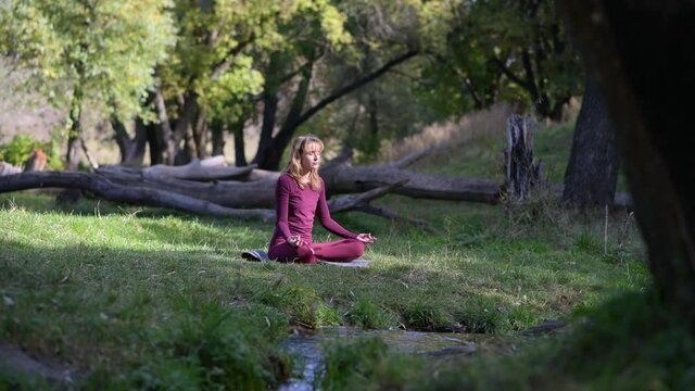 Woman sit static in yoga lotus pose in peaceful forest with woods, light wind and flowing water stream