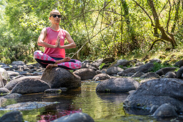 Mujer joven y atractiva haciendo yoga en un parque natural al aire libre con rio, arboles y rocas