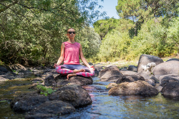 Fototapeta na wymiar Mujer joven y atractiva haciendo yoga en un parque natural al aire libre con rio, arboles y rocas