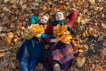 Mom and daughter lie on autumn leaves in the park and hold a bouquet of yellow leaves in their hand.