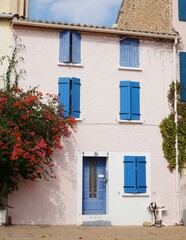 Fototapeta na wymiar House front adorbed with red flowers in bloom and featuring blue door and window shutters in Mediterranean seaside village of Leucate, southern France 