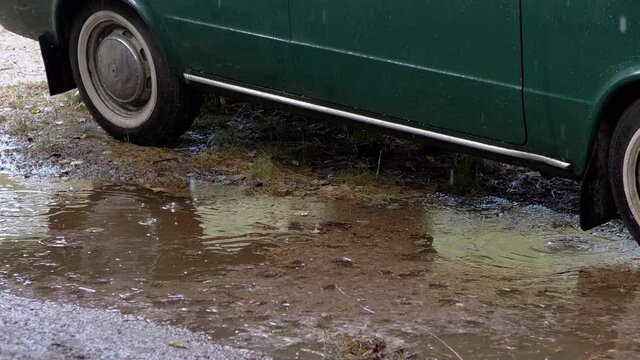 Rain Drops Dripping into a Muddy Puddle Near a Parked Old Car in Nature in Wood. Wheels of a green vintage car got stucked in the mud in the rain. Dirt, slush, rainy weather outdoors. Trip. 4K.