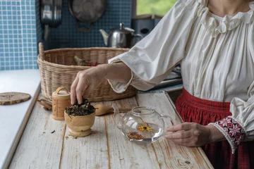 Foto op Canvas A girl in a folk costume makes tea from herbs and flowers. A woman in the kitchen is preparing her own tea. © Sergei