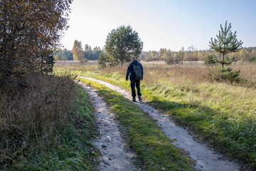 Obraz premium A tourist walks along a country road early in the morning. A man with a backpack on the background of a field illuminated by sunlight.
