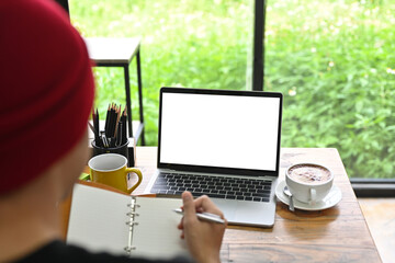 Behind view of a creative man taking notes while sitting at the wooden working desk surrounded by a...