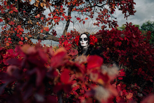 Woman with sugar skull makeup and red roses dressed as Santa Muerte is against background of autumn forest.