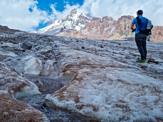 A man standing at the foothill of Gergeti Glacier, with the view on Mount Kazbeg, Caucasus, Georgia. Barren slopes below the snow-capped peak. Few clouds gathering around the sharp peak. Tranquillity