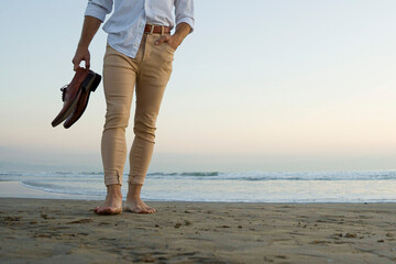  Classy elegant man in white shirt and beige pants walking on the beach with his shoes in hands