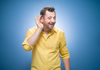 Half length man holding palm near his ear trying to listen something interesting over blue...