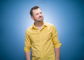Strict young man thinks. Portrait of doubtful guy with confusion face expression over blue background, dresses in yellow shirt. Careful man, copy space for text