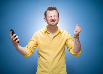 Irritated young man talks on the phone over blue background, dresses in yellow shirt. Annoyed guy talking. Stressed person using smartphone